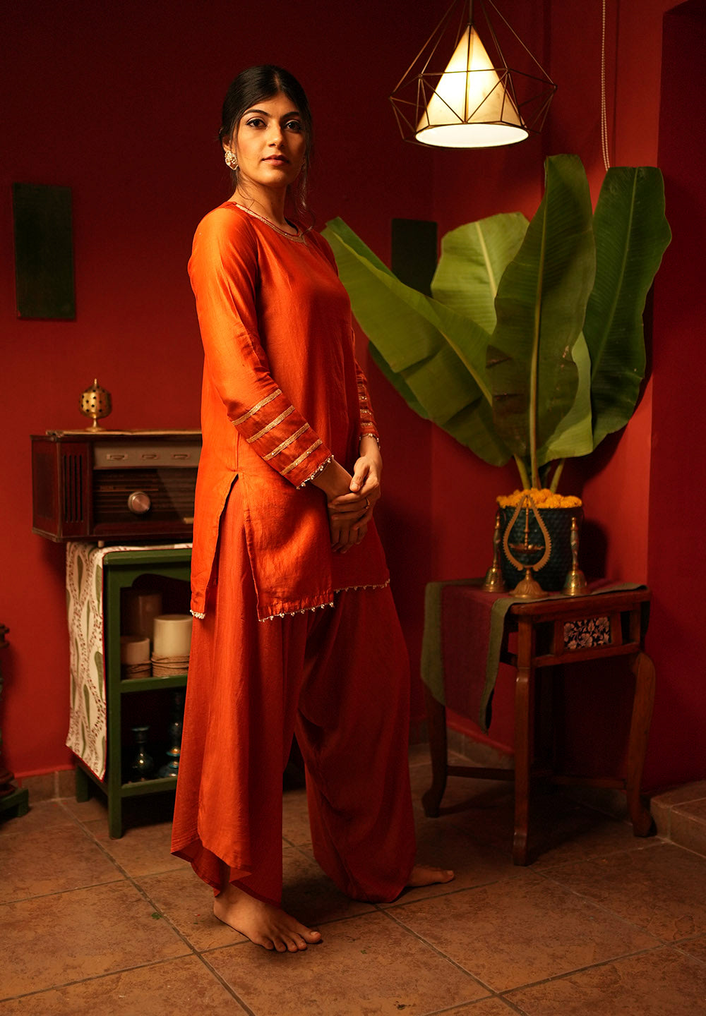 Woman in traditional orange outfit standing in a room with red walls and decorative plants.